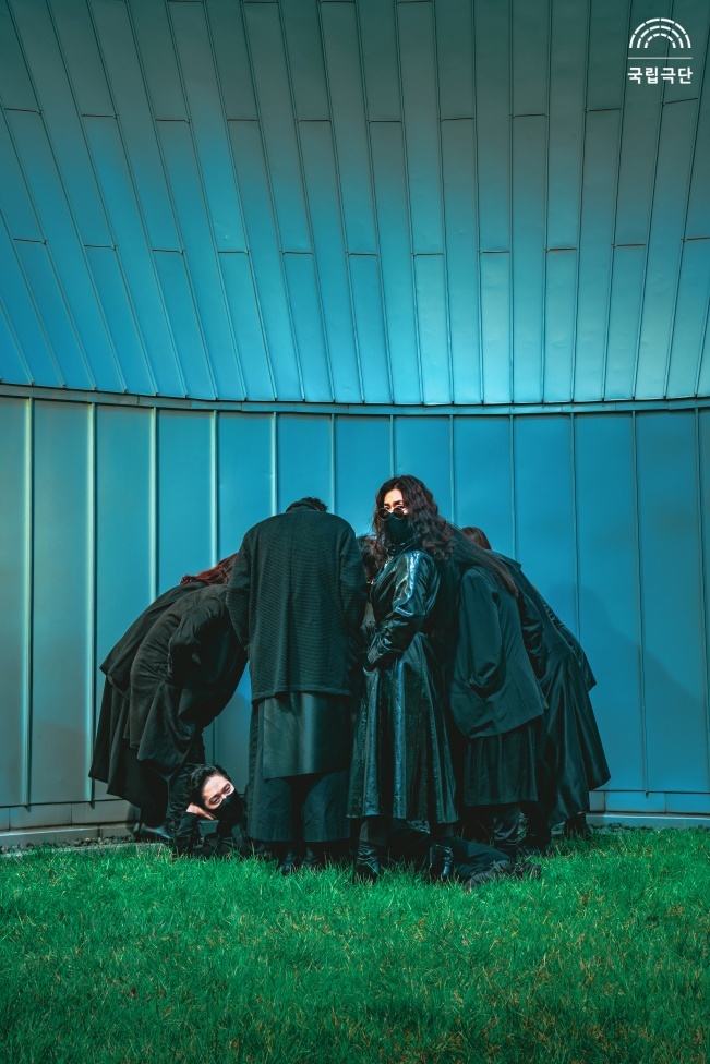 공연 홍보사진. 10명의 배우가 검은색 의상을 입고 둥글게 둘러서 있습니다. 그 중 두 명은 정면을 바라보고 있습니다.